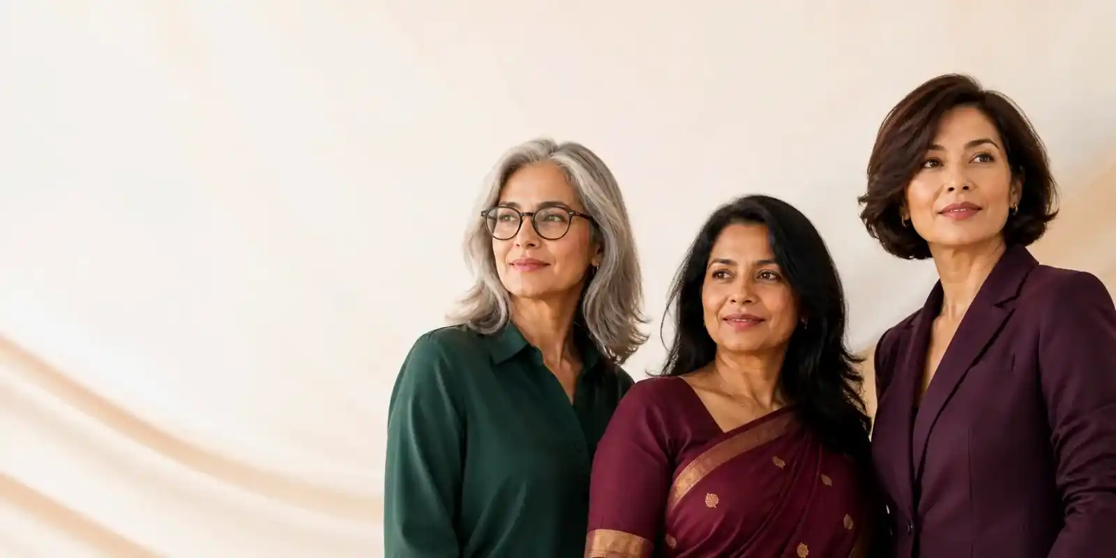 Three women standing together against a warm studio backdrop, one in a green shirt and glasses, one in a plum saree, one in a plum jacket
